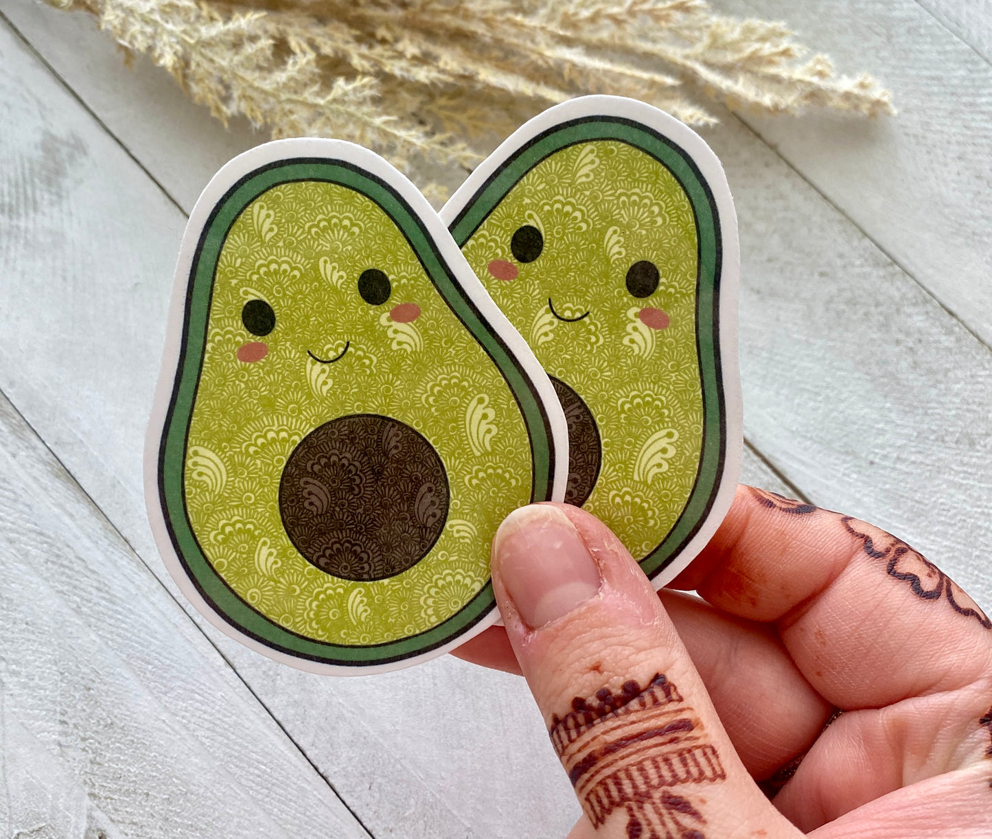 Two avocado stickers with faces held by a hand on a light wooden surface.