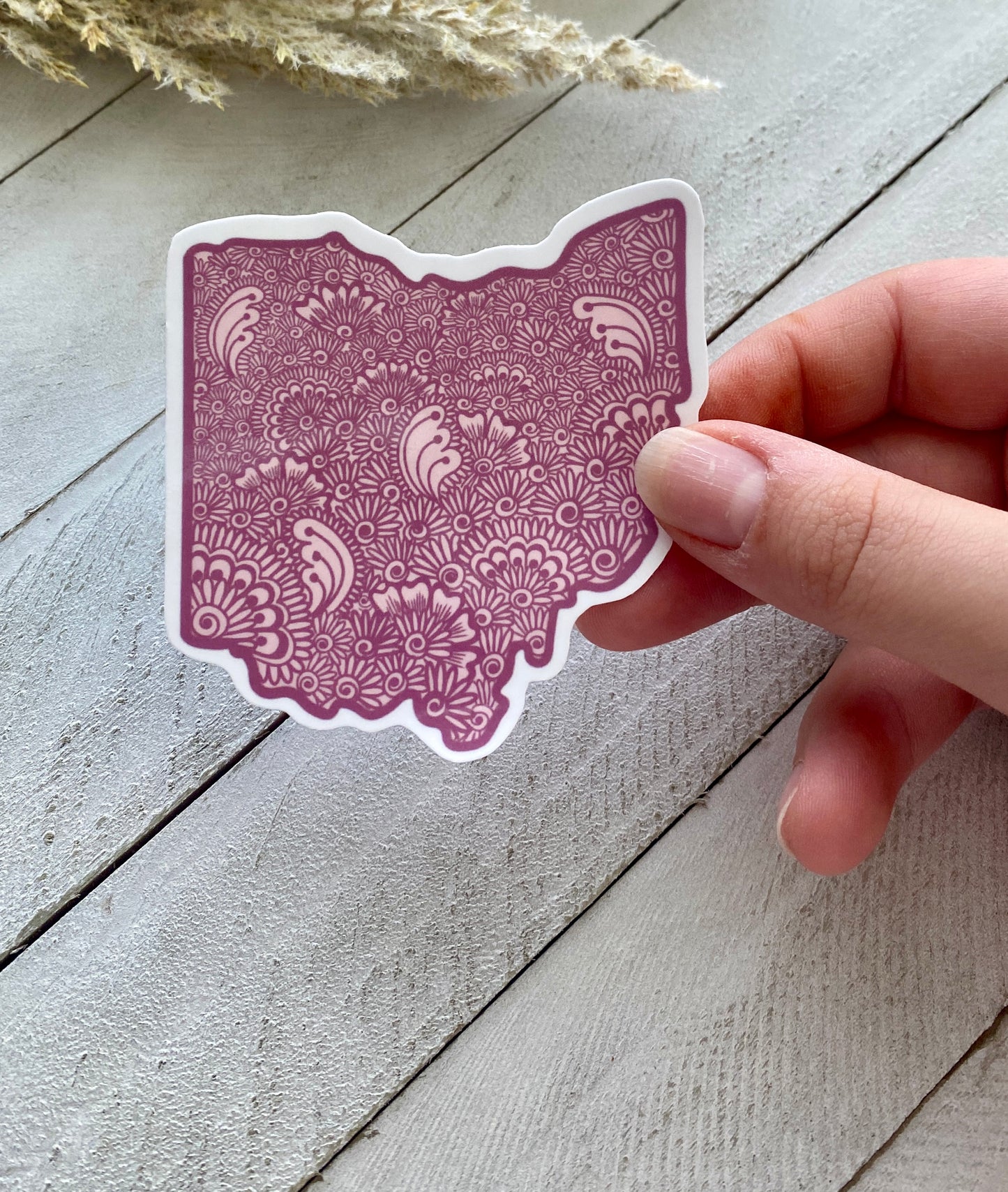 A hand holding a purple Ohio State sticker with a detailed henna design over a wood background