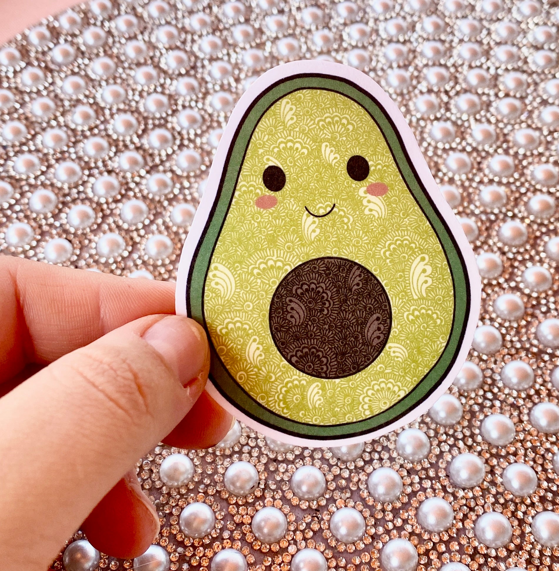 Sticker of a cute avocado design held by a hand on a decorative background with pearls.