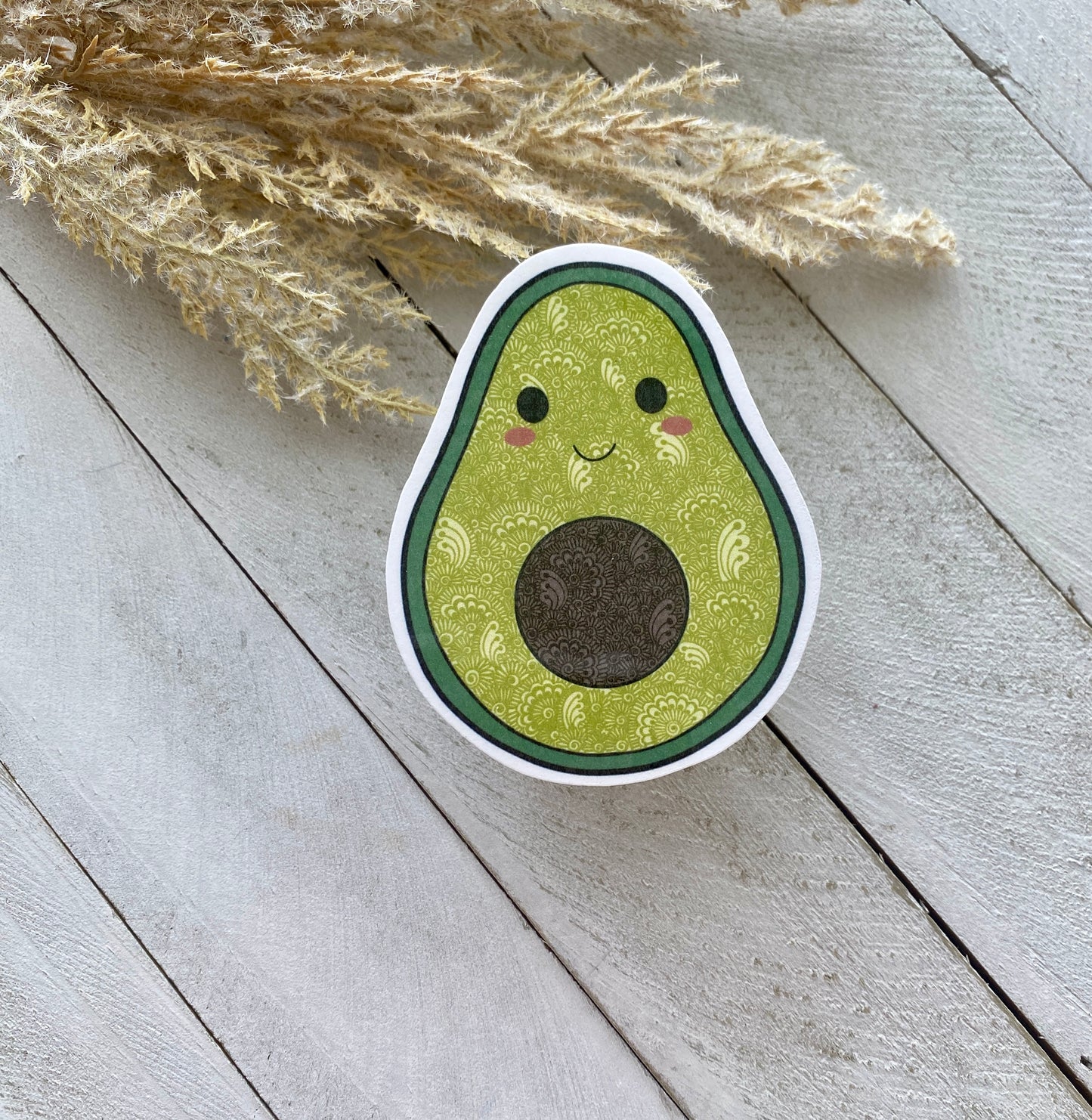 Sticker of a smiling avocado on a wooden surface with dried plants.