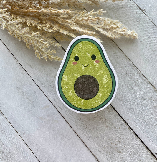 Sticker of a smiling avocado on a wooden surface with dried plants.