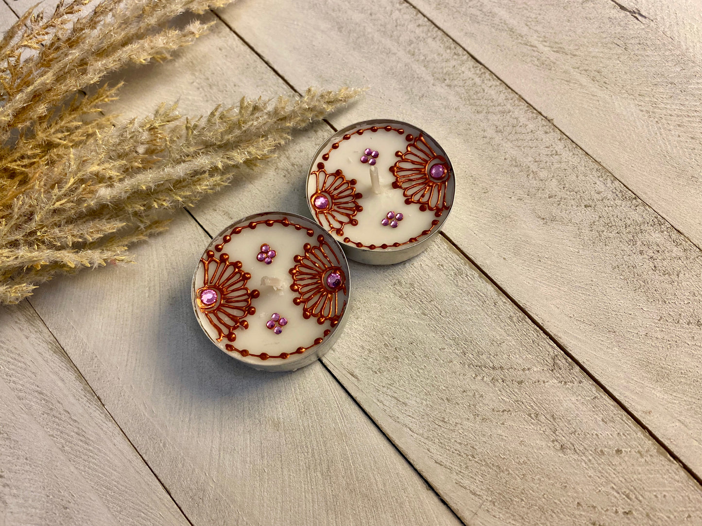 Two tea light with bronze painted florals each with pink gemstones sitting against a wood surface. 
