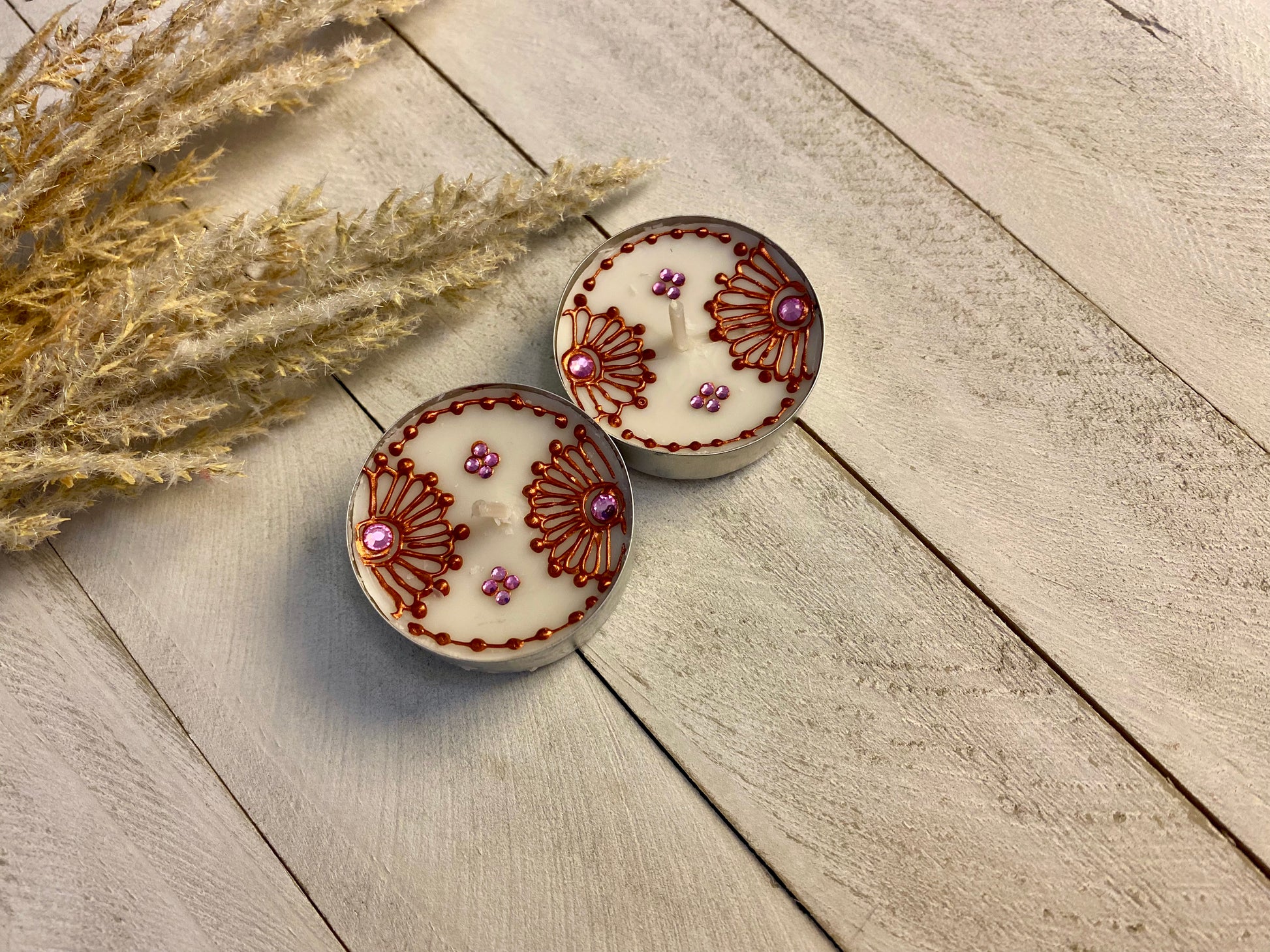 Two tea light with bronze painted florals each with pink gemstones sitting against a wood surface. 