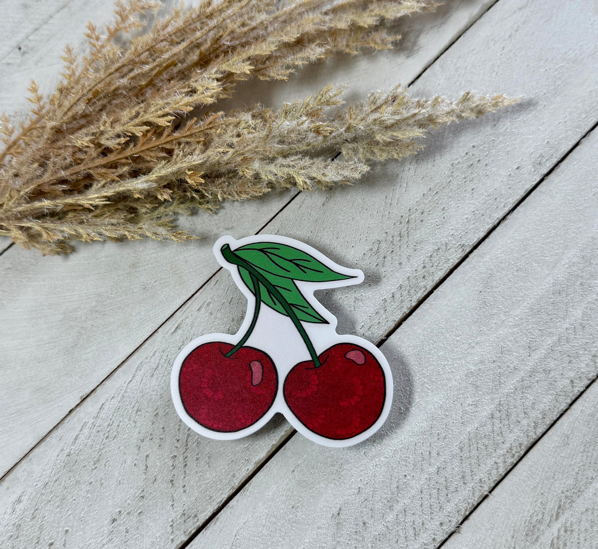 A sticker depicting two red cherries with a green leaf on top, placed on a wooden surface with straw in the background.
