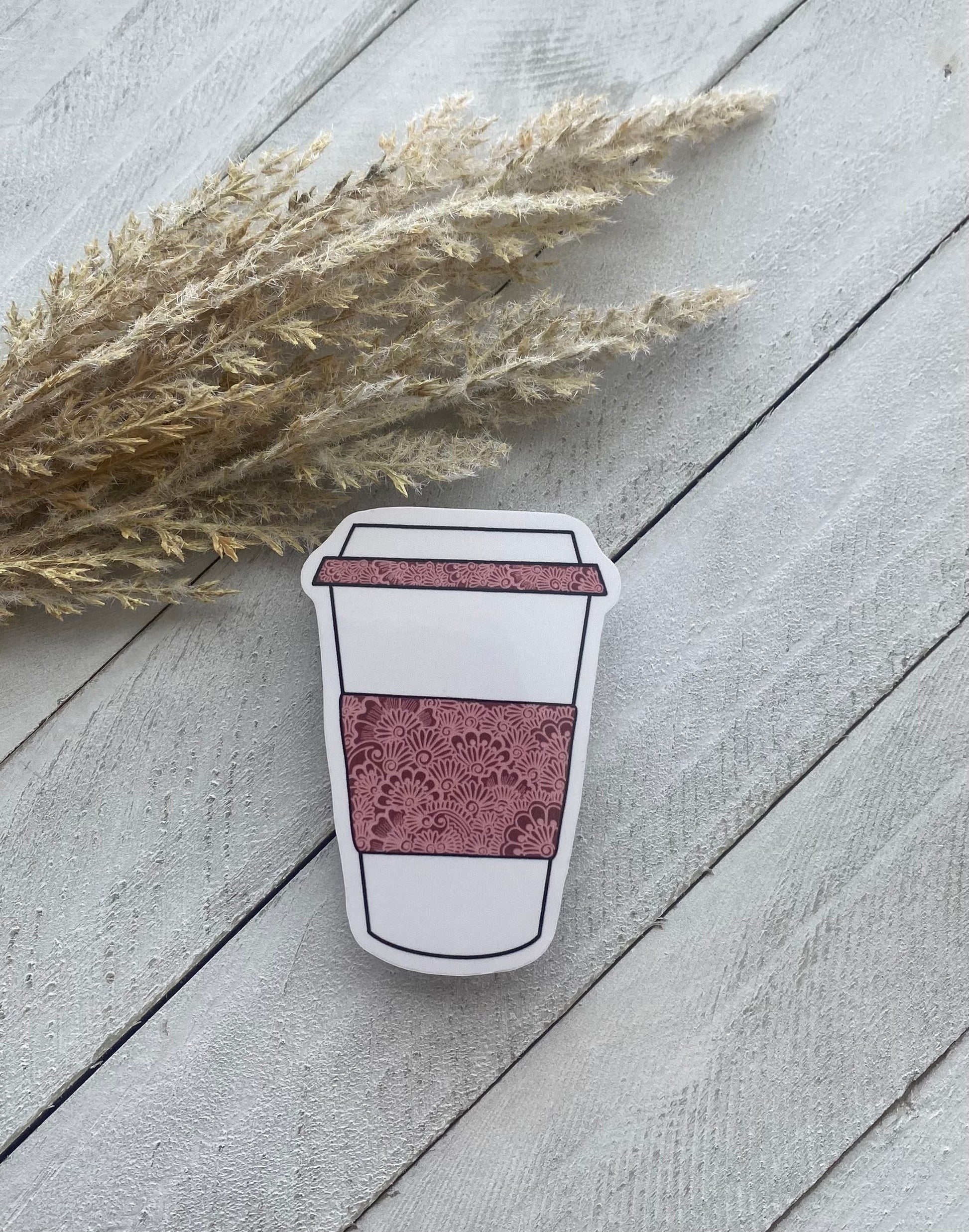 A sticker with a coffee cup design, laminated and placed on a wooden surface, with dried plant stems to the side.