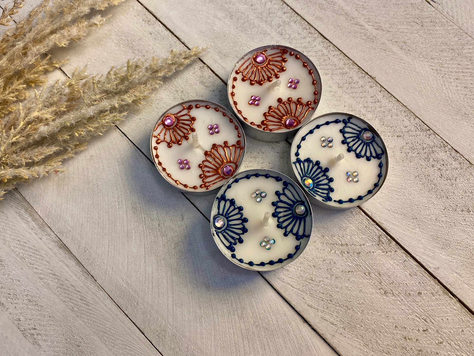 Four hand-painted tea lights with floral patterns and rhinestone embellishments, displayed on a wooden surface with dried wheat in the background.