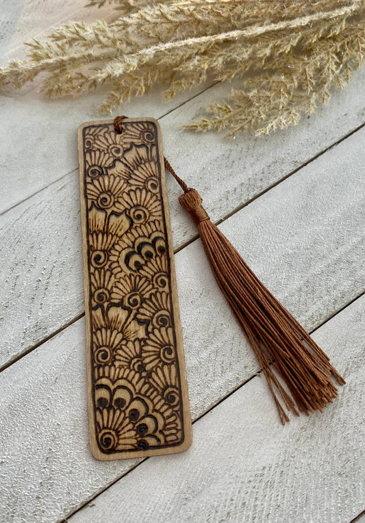 A bookmark with a floral henna design, light brown stained wood, and a tassel attached.