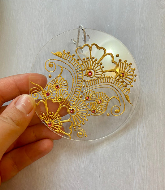 A hand holding a clear circle acrylic ornament with a gold henna designs and pink rhinestones