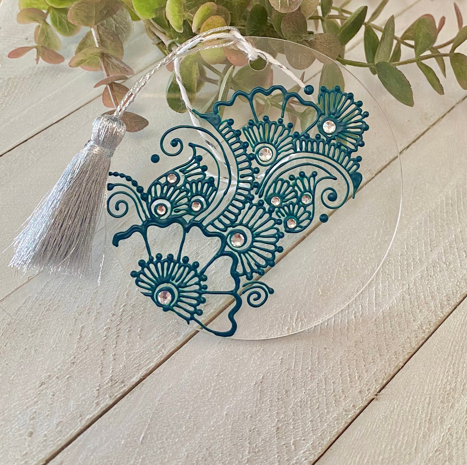 A clear circle acrylic ornament with a green henna design and clear rhinestones and a silver tassel against a wood background with greenery 
