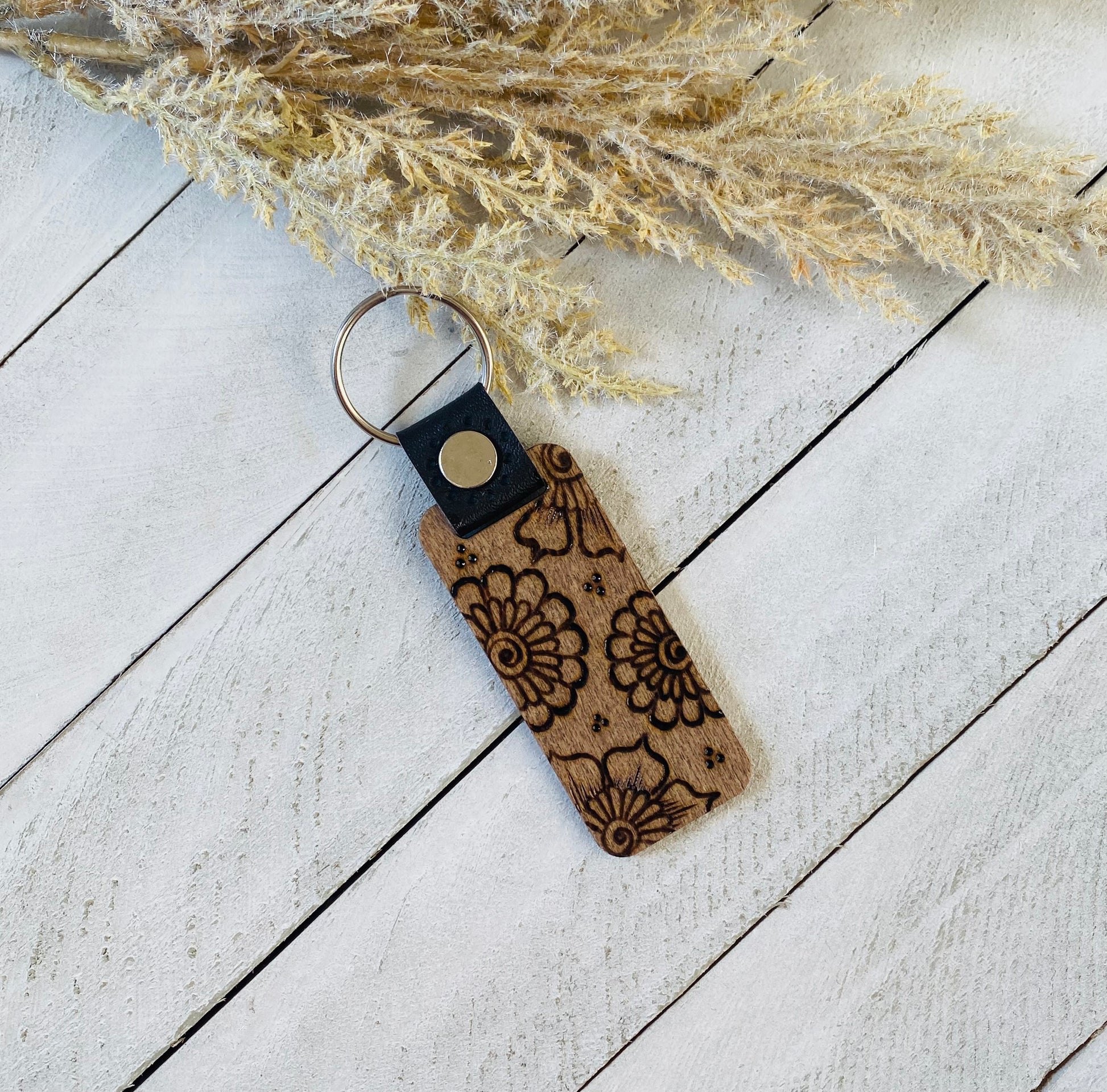 A wood burned keychain with a floral henna design, placed on a white wooden surface, next to some dried plant stems.
