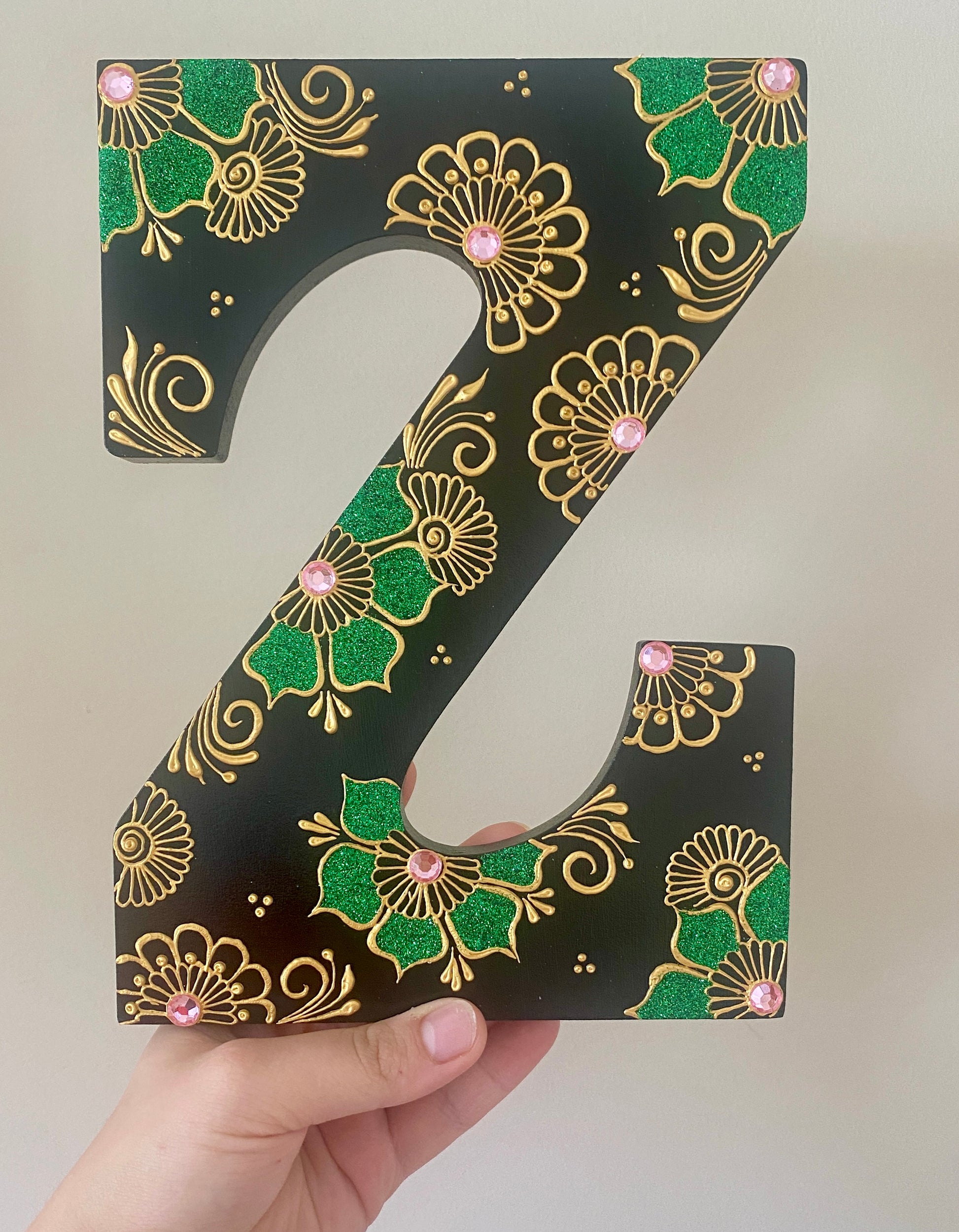 A hand holding a black letter Z with gold painted florals with pink rhinestones and green glitter 