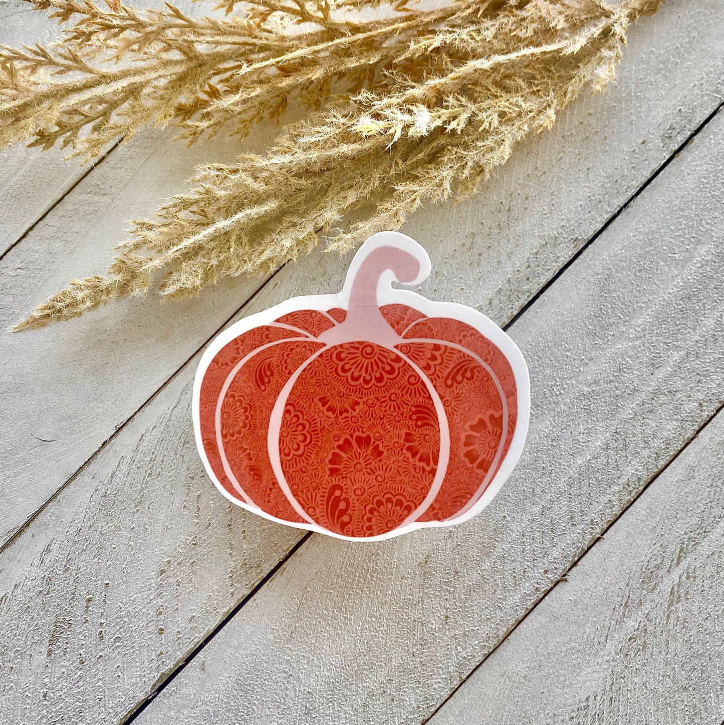 A sticker with a pumpkin design placed on a wooden surface, with dried plant stems in the background.