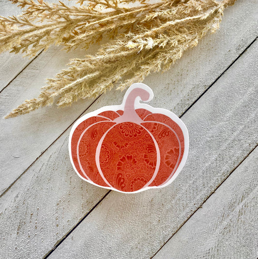 A sticker with a pumpkin design placed on a wooden surface, with dried plant stems in the background.