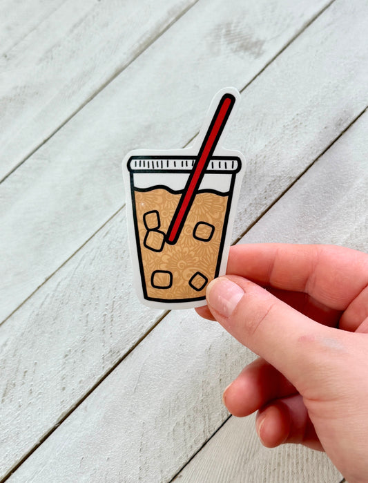 Sticker of a coffee cup with a straw held by a hand on a light wooden surface
