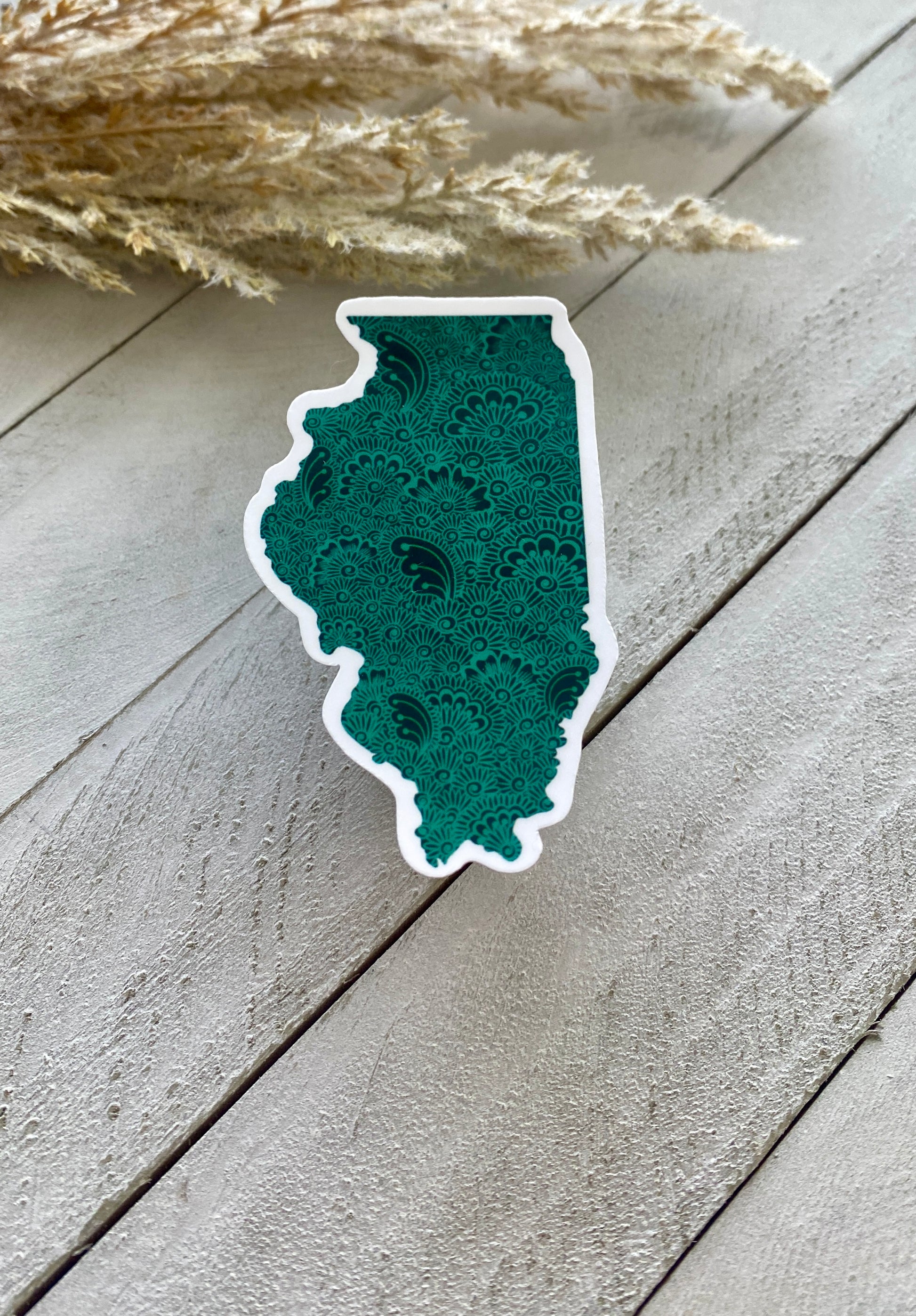 An Illinois state sticker with a green background and henna designs inside placed on a white wooden surface