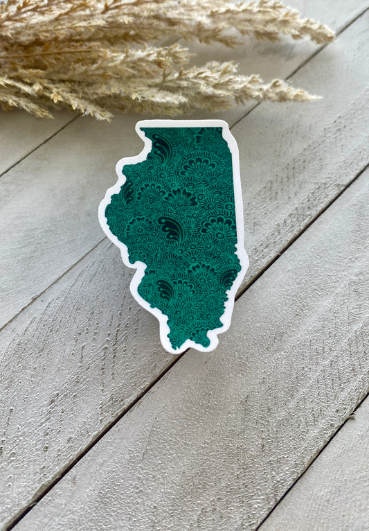 An Illinois state sticker with a green background and henna designs inside placed on a white wooden surface