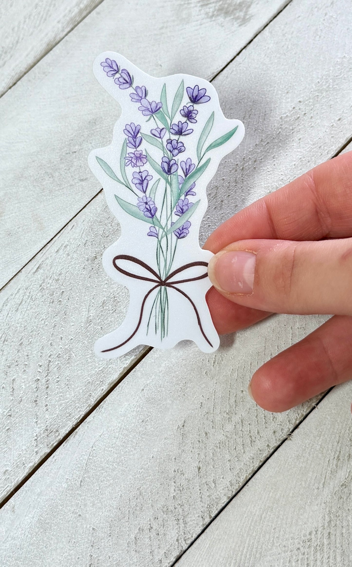 Hand holding a sticker with a design of lavender flowers and a brown bow on a light wooden surface.