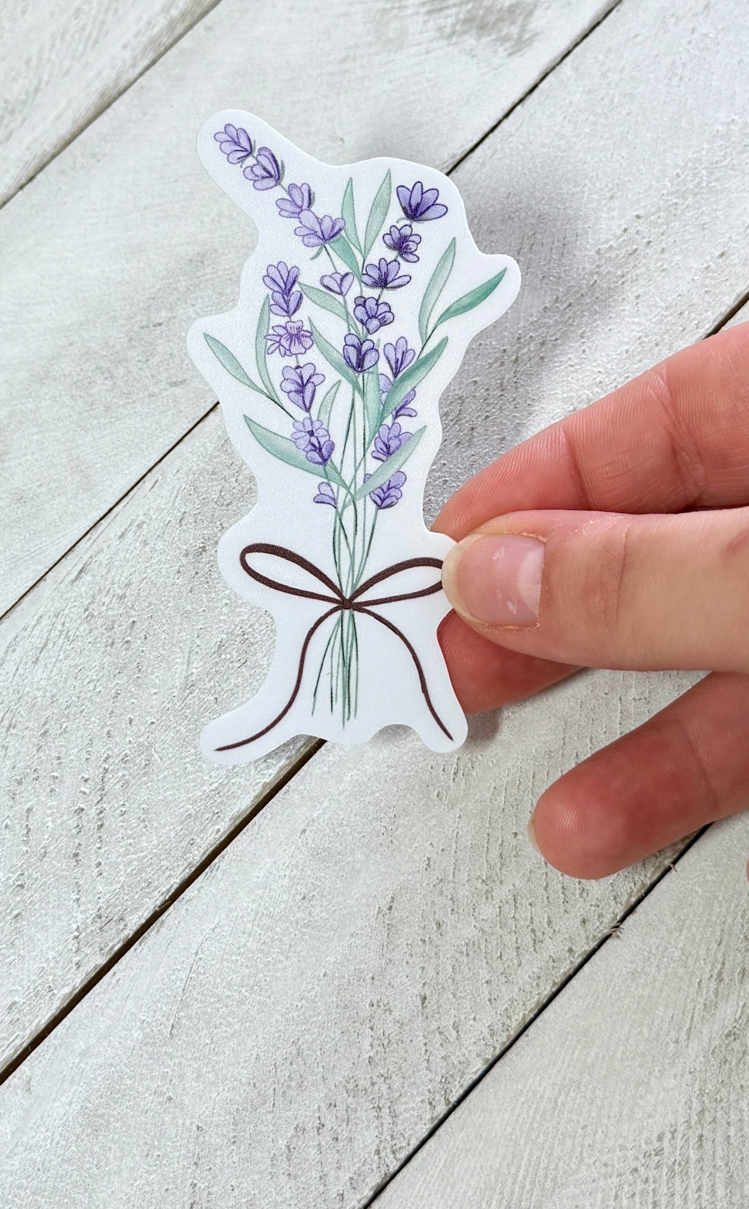 Hand holding a sticker with a design of lavender flowers and a brown bow on a light wooden surface.
