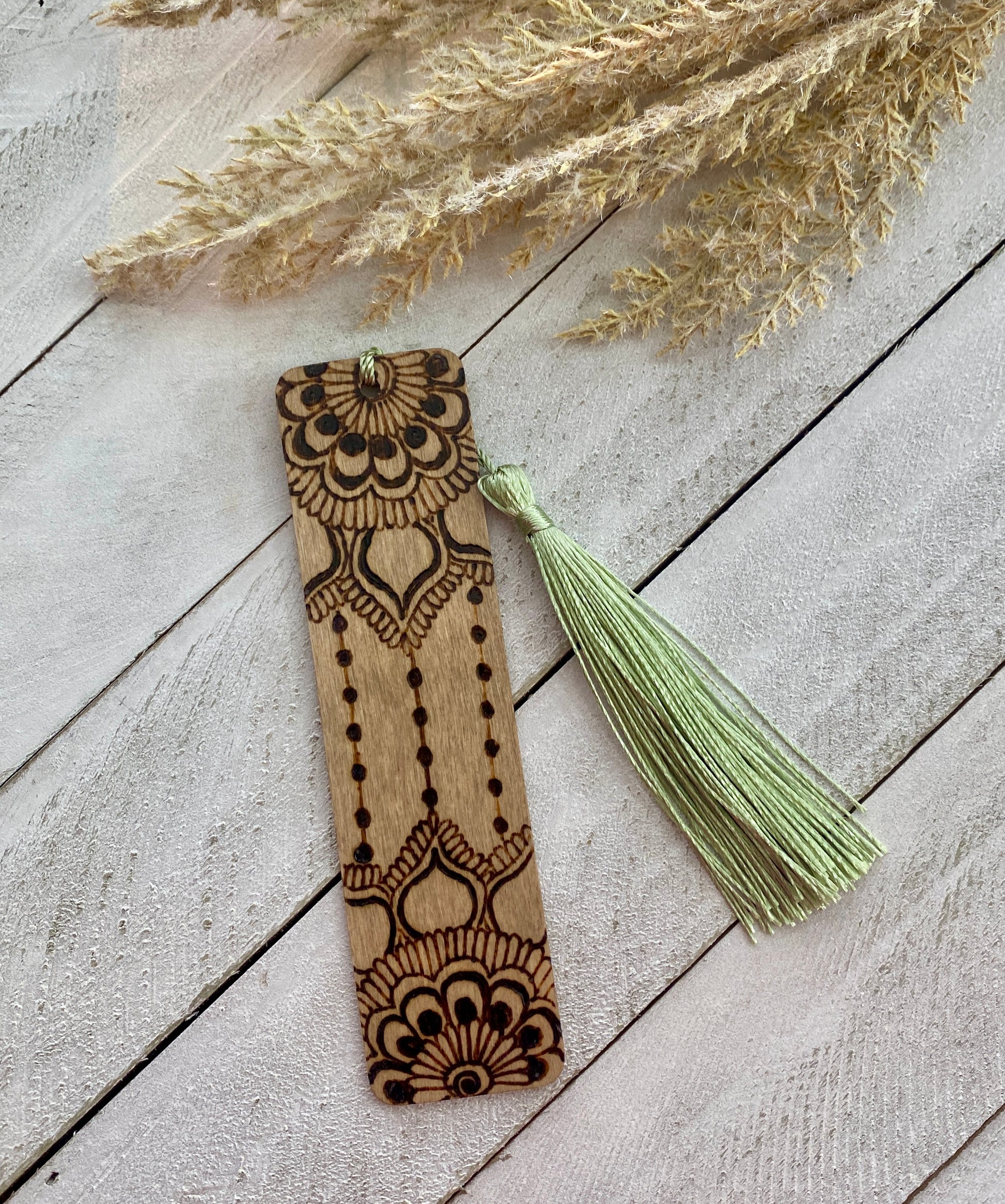 A bookmark with a floral henna design, stained light brown, placed on a white surface with dried plant stems in the background.