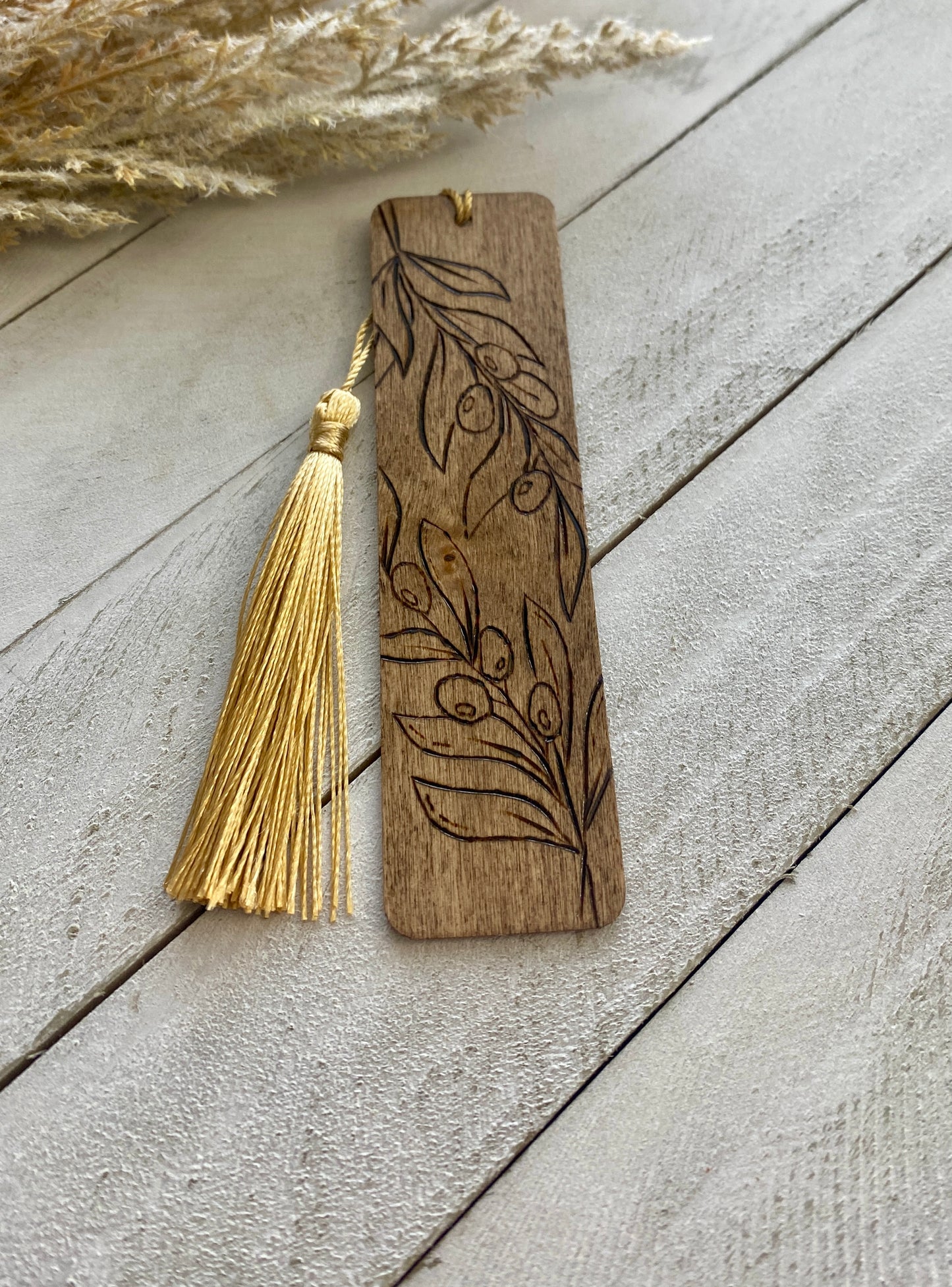 A bookmark with a dark walnut color finish, featuring an intricate henna design of olive branches, accompanied by a tassel with golden strands.