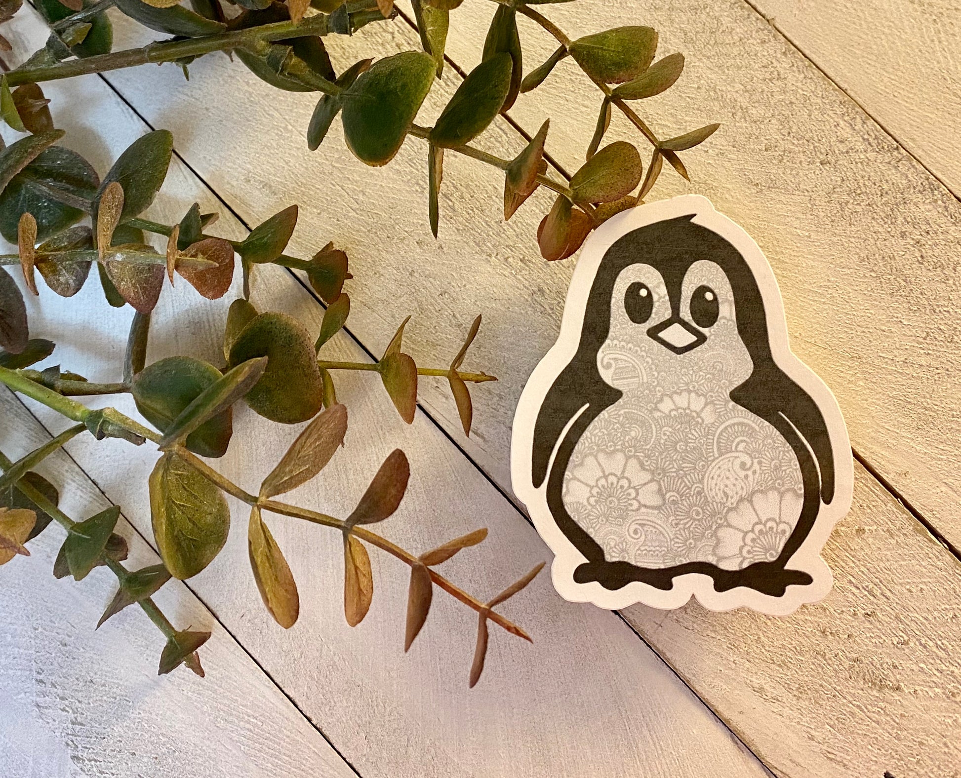 A sticker of a hand-drawn penguin with a gray background, placed on a white surface surrounded by green plant leaves.