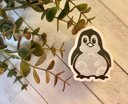 A sticker of a hand-drawn penguin with a gray background, placed on a white surface surrounded by green plant leaves.