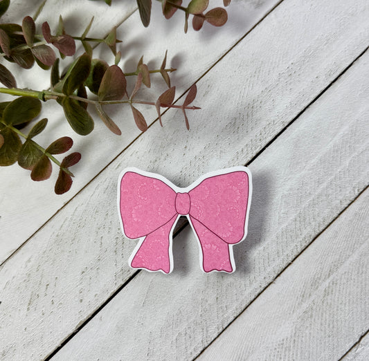 Pink bow-shaped sticker on a light wooden surface with green leaves.