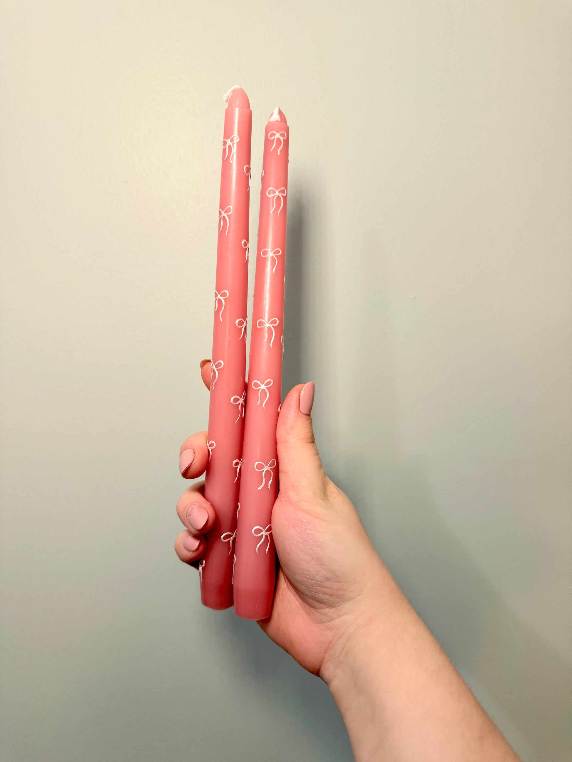 Hand holding two pink tapered candles with white patterns against a plain background