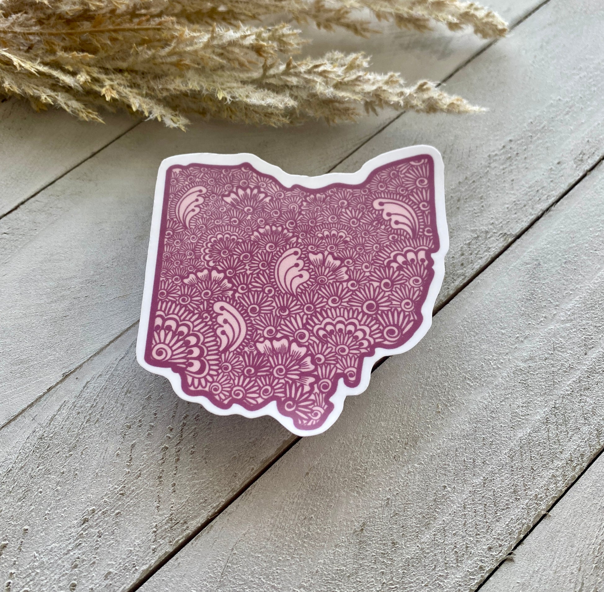 A sticker with a purple background and a detailed floral pattern shaped like the state of Ohio, placed on a white wooden surface with a field of dry grasses in the background.