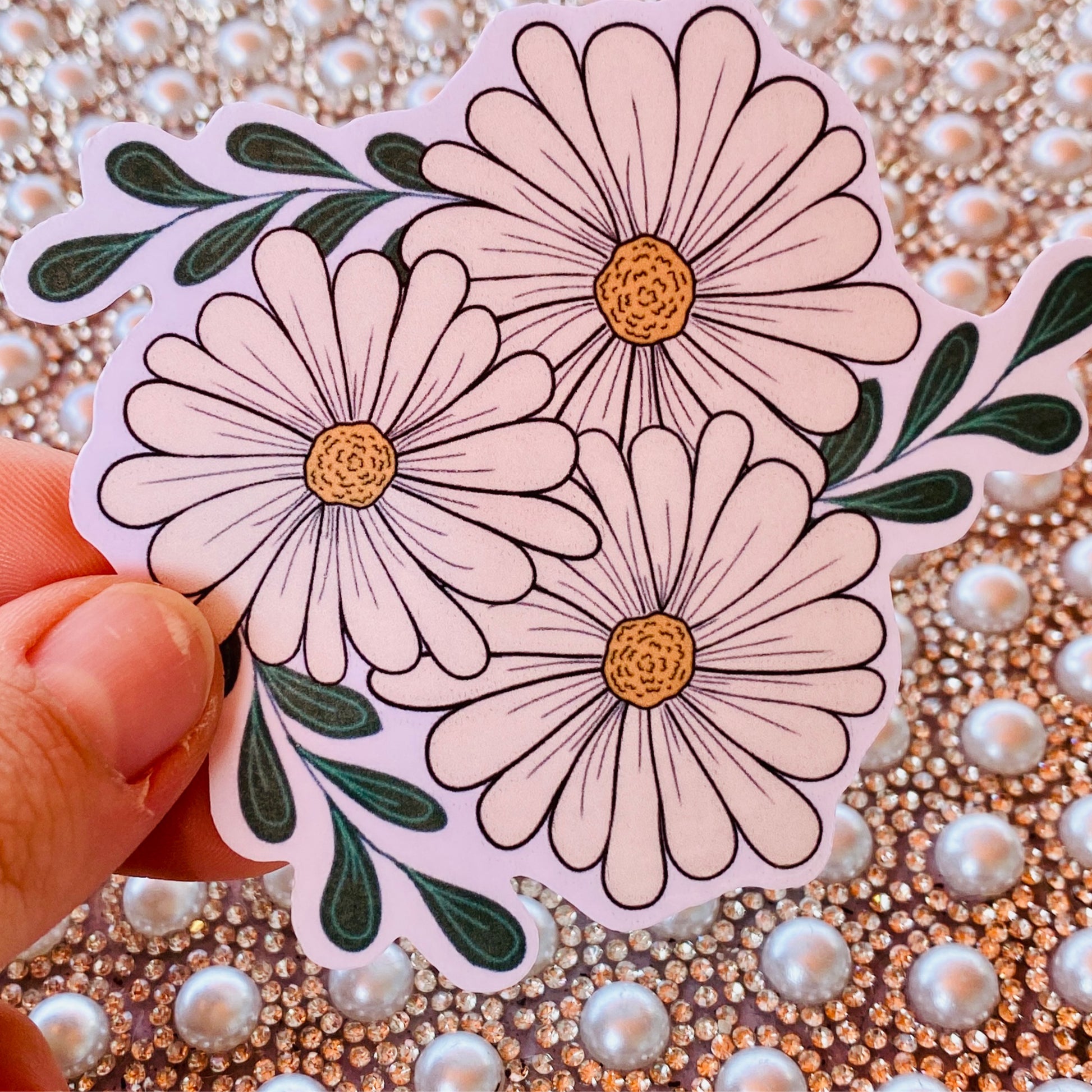 A hand holding a sticker with three daisy flowers against a sparkle background