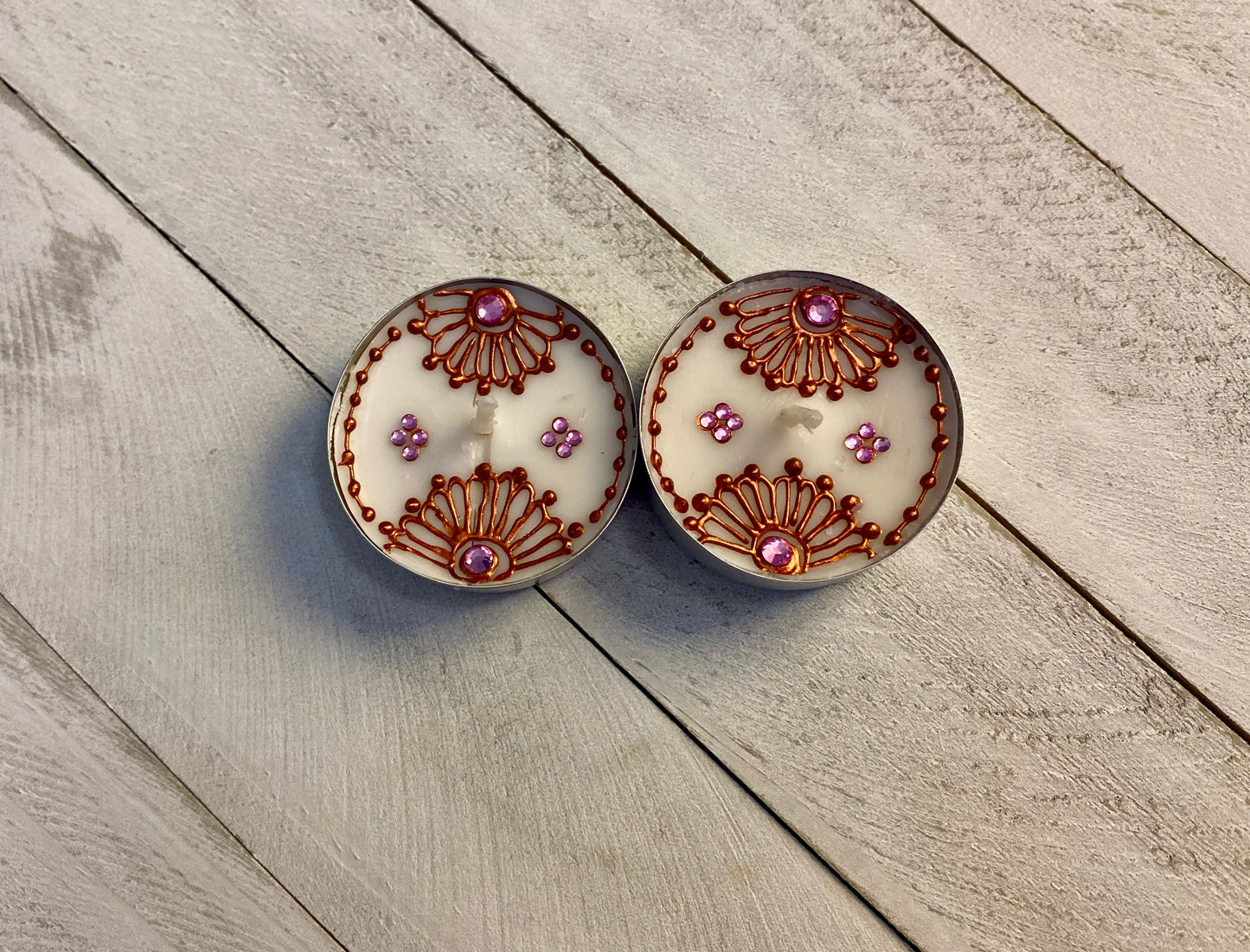 Two tea lights featuring bronze painted florals with pink rhinestones against a wood background 
