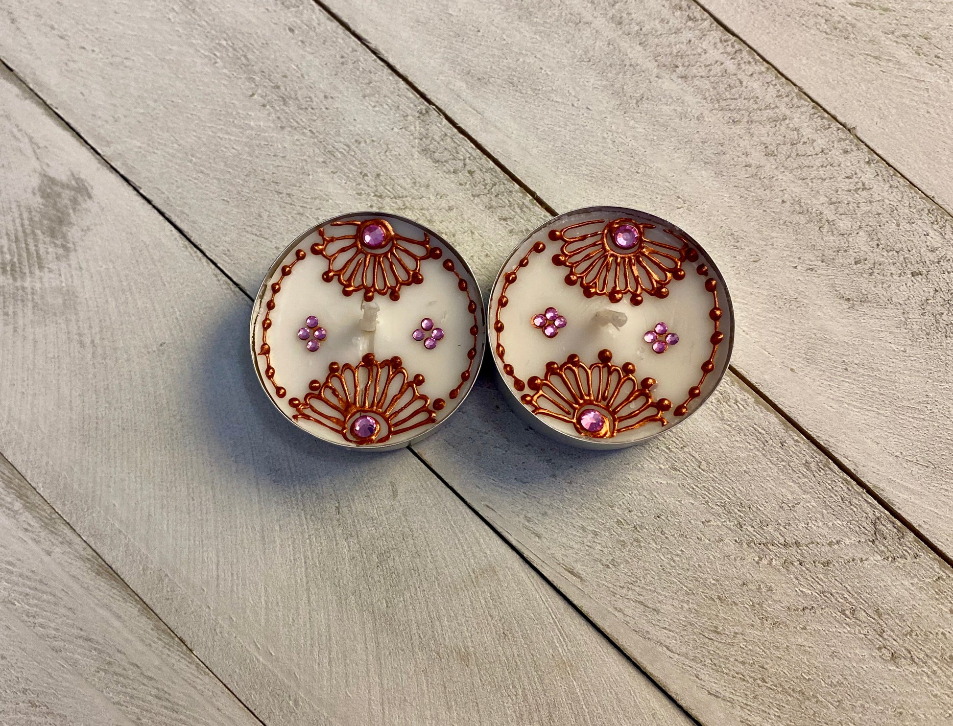 Two tea lights featuring bronze painted florals with pink rhinestones against a wood background 