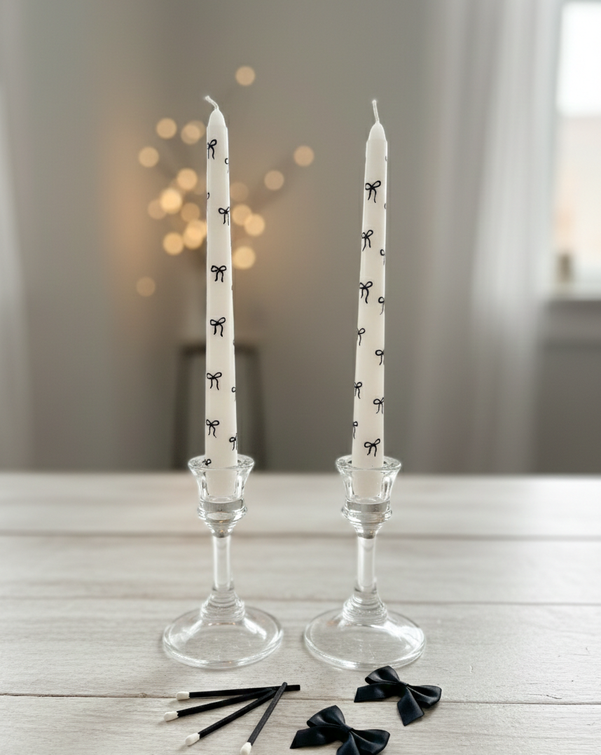 Two white candles with black bow patterns in glass candlesticks on a wooden surface.