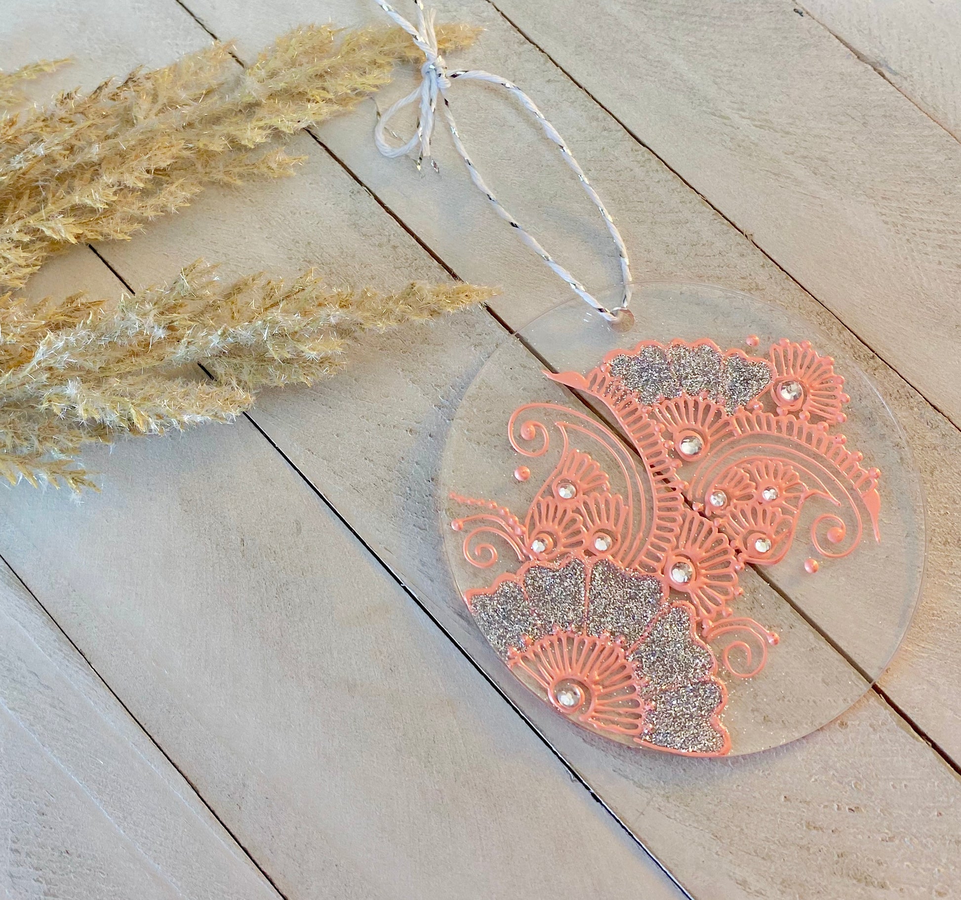 Circle clear acrylic ornament with pink henna designs , silver glitter and rhinestones against a wood background 